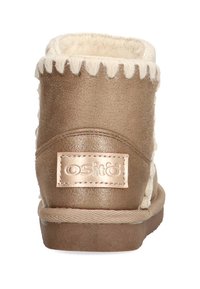 Botín marrón hasta el tobillo con un forro suave y esponjoso, costuras decorativas y un parche de logo metálico. Suela plana con un acabado texturizado.