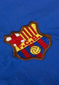 FC Barcelona geborduurd embleem op een blauwe stof achtergrond, met rode en gele velden, golvende lijnen en een cirkelvormige voetbaldetail.