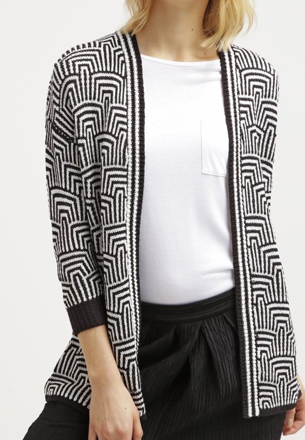 Černobíle vzorovaný kardigan s geometrickými vzory, otevřený vpředu a s žebrovanými manžetami. Nosený přes jednoduchý bílý top a černou skládací sukni.