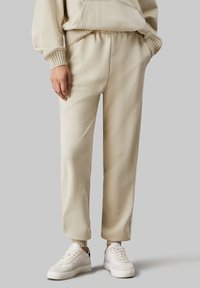 Pantalones de chándal color crema con puños y cinturilla elásticos, fabricados en una tela suave. Combinados con zapatillas blancas con acentos texturizados.