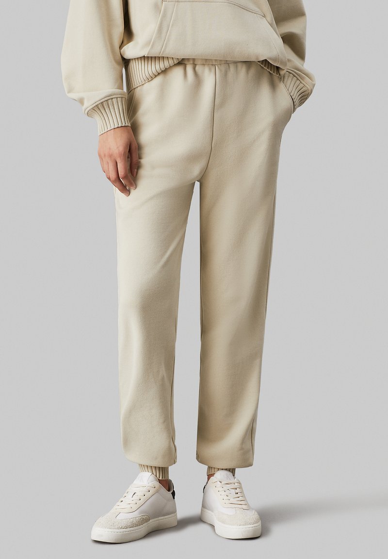 Pantalones de chándal color crema con puños y cinturilla elásticos, fabricados en una tela suave. Combinados con zapatillas blancas con acentos texturizados.