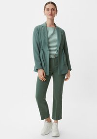 Helles grünes Blazer mit Zugband in der Taille über einem gestreiften Oberteil. Passende grüne Hose mit gerader Passform und weißen Sneakern. Einfaches, modernes Design.