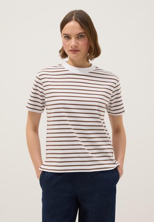 Jonge vrouw met kort haar, gekleed in een wit T-shirt met bruine horizontale strepen en een donkere broek, staand met haar handen in de zakken.