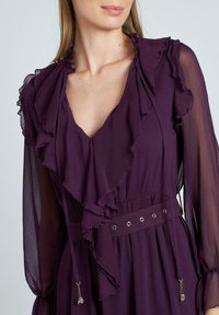 Vestido púrpura con cuello acampanado, mangas largas transparentes y una cintura ajustada con ojales decorativos y acentos de cordón.