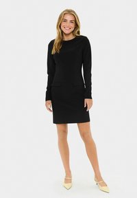 Femme souriante debout, portant une robe noire ajustée à manches longues avec des poches à l'avant et des talons bas jaunes pâles, sur fond blanc.