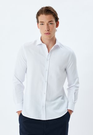 Camisa blanca de manga larga y botón con una textura suave, presenta un cuello puntiagudo y un dobladillo redondeado, combinada con pantalones azul marino.