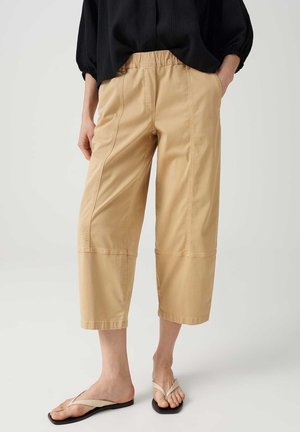 Person trägt beige, weit geschnittene, knielange Hosen mit elastischem Bund, schwarzes Top und hellbeige Sandalen auf hellem Hintergrund.