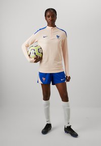 Nike moterų futbolo apranga, sudaranti šviesiai rožinės spalvos ilgomis rankovėmis viršutinius drabužius su mėlynu apykakle ir logotipu, mėlynos šortai su emblema ir juodi sportbačiai.