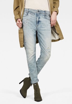 Light blue denim jeans with a relaxed fit, distressed details, and patchwork. Paired with olive-green ankle boots and a beige top.