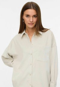 Young woman with straight brown hair wearing a loose light beige button-up shirt and dangling pearl earrings, against a plain white background.