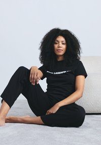 Tricou negru din bumbac cu text alb, asortat cu pantaloni negri. Modelul are păr creț și este așezat pe o suprafață texturată deschisă la culoare.