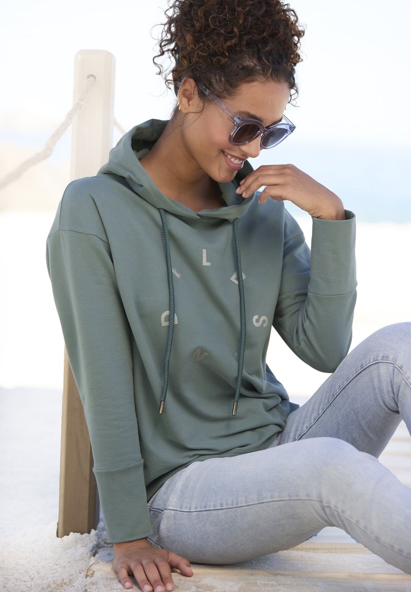 Felpa verde con cappuccio e cordini, dal tessuto morbido, caratterizzata da una stampa minimalista. Abbinata a jeans skinny blu chiaro.