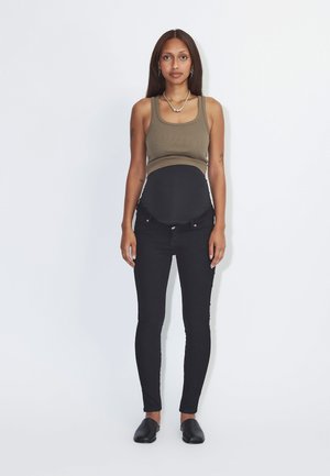 Mulher a usar camisola de alças verde-oliva, jeans de gravidez pretos e sapatos pretos slip-on, de pé contra um fundo branco.