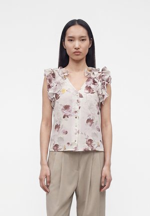 Kobieta nosząca bezrękawnikową kwiecistą bluzkę z falbanami na ramionach oraz beżowe spodnie, stojąca na jednolitym białym tle.