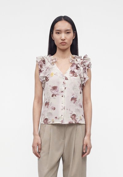 Femme portant un chemisier sans manches à motifs floraux avec des épaules volantées et un pantalon beige, debout devant un fond blanc uni.
