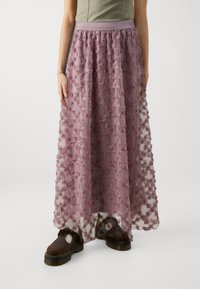 Lange, doorschijnende rok in mauve met 3D bloemversieringen, voorzien van een aansluitende tailleband en gecombineerd met bruine plateau-schoenen.