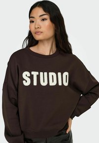 Bruine sweatshirt met witte "STUDIO" tekst, ronde hals en verlaagde schouders, gemaakt van zachte, gemêleerde stof.