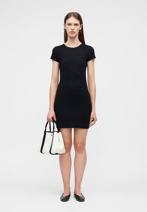 Γυναίκα με εφαρμοστό μαύρο φόρεμα κοντού μανικιού, κρατώντας μια λευκή τσάντα με μαύρα χερούλια, φορώντας μαύρα επίπεδα παπούτσια, στέκεται σε απλό φόντο.