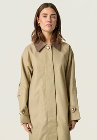 Beige színű trench coat barna velúrbőr gallérral, gombos záródással és széles ujjakkal. Sima anyag, minimál stílusú design, látható minták nélkül.
