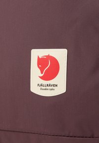 Parche del logo de Fjällräven sobre tela de color púrpura oscuro, con un rectángulo blanco que presenta la silueta de un zorro rojo y el nombre de la marca y el año debajo.