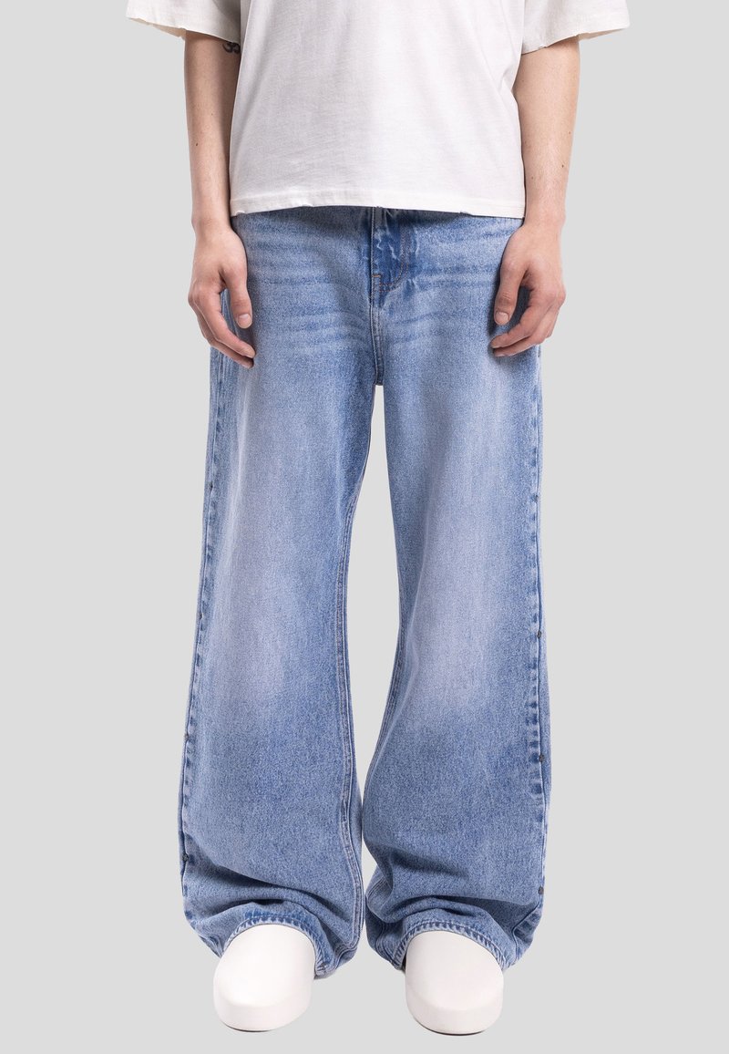 Personne portant un jean large bleu clair, un t-shirt blanc oversize et des chaussures blanches, debout devant un fond gris uni.