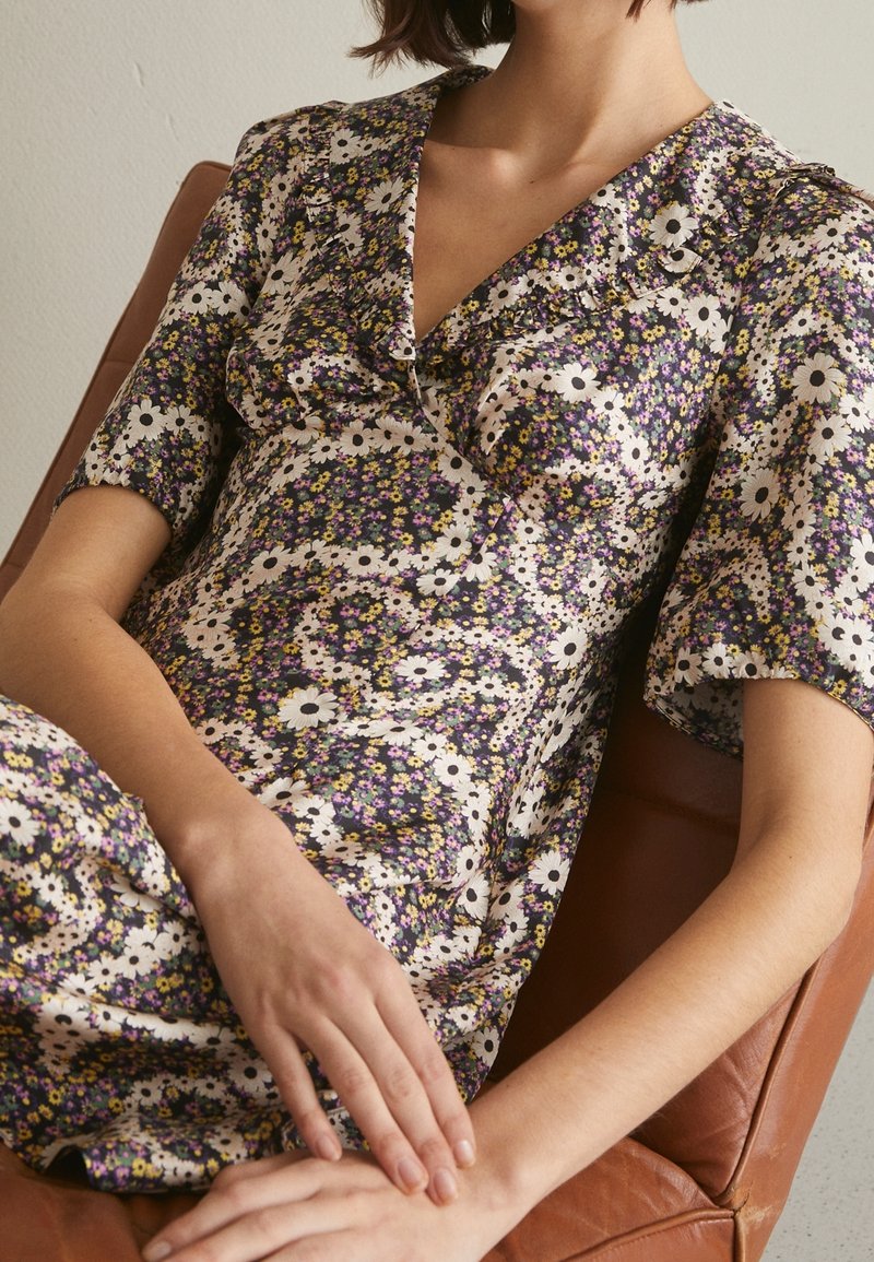 Robe fleurie en tissu léger arborant un motif multicolore avec des fleurs violettes, blanches et jaunes. Encolure en V et détails à volants.