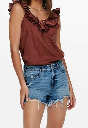 Nainen, jolla on päällään hihaton ruskea röyhelöinen paita ja korkeavyötäröiset kulutetut siniset farkkushortsit, joissa niittivyö.