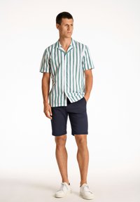 Raitapaitainen lyhythihainen paita vihreän ja valkoisen sävyissä, yhdistettynä laivastonsinisiin shortsit. Mallilla on päällään valkoiset lenkkarit, mikä luo rentoa kesätyyliä.