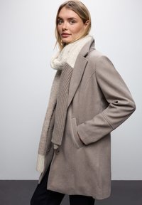 Manteau en laine gris clair avec un design structuré, de grands revers et des poches latérales. Associé à une écharpe en tricot crème texturée.