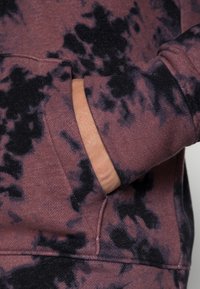 Sudadera con capucha en malva oscuro con patrón de tie-dye negro. Presenta un bolsillo canguro y puños acanalados, fabricada en un tejido suave y texturizado.
