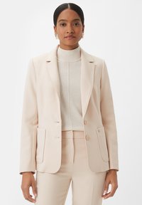 Blazer tailored di colore rosa chiaro con rever a punta, chiusura con un solo bottone e due tasche frontali. Indossato sopra un maglione beige a collo alto.