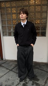 Giovane con capelli corti che indossa un maglione nero, una camicia bianca con colletto e pantaloni larghi scuri, in piedi con le mani nelle tasche su un pavimento piastrellato.