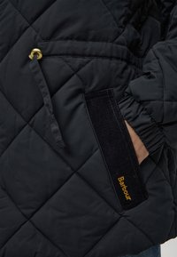 Main dans la poche d'une veste matelassée noire Barbour avec poignet élastique et cordon de serrage avec détail en métal doré.