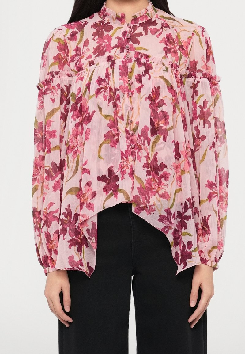 Femme portant une blouse fleurie rose transparente avec de longues manches bouffantes, des détails en volants et des boutons sur le devant, associée à un pantalon noir.