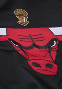 Schwarzer Stoff mit einem großen roten gestickten Chicago Bulls-Logo mit schwarzen Akzenten und einem goldenen Basketball-Trophäen-Logo darüber.