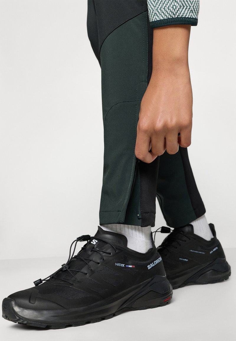 Czarne buty sportowe z teksturowanym wzorem, z systemem sznurowania i grubą podeszwą. Noszone z zielonymi spodniami sportowymi i białymi skarpetkami.