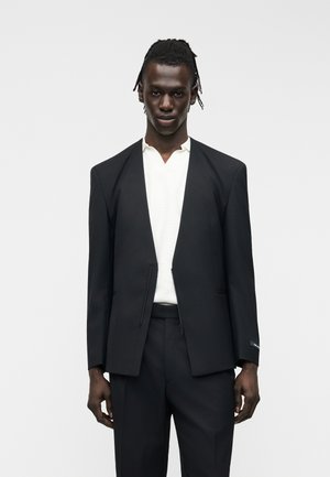 Ψηλός άνθρωπος με σκούρο δέρμα και πλεξούδες που φοράει μαύρο σακάκι πάνω από λευκό πουκάμισο και μαύρο παντελόνι, στέκεται μπροστά σε ένα απλό φόντο.