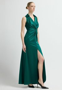 Szmaragdowo zielona sukienka kopertowa z głębokim dekoltem w serek, bez rękawów, z wiązaniem w pasie i wysokim rozcięciem na udzie. Gładkie, satynowe wykończenie.