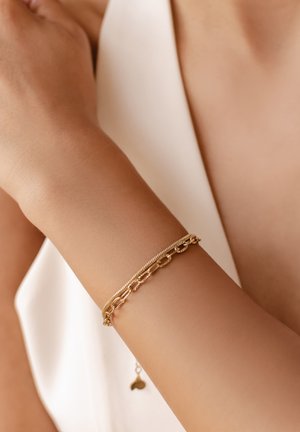 Goldarmband mit einer Mischung aus verknüpfter Kette und strukturierten Elementen, am Unterarm getragen, mit einem glatten Hautton und einem subtilen, hängenden Charm.
