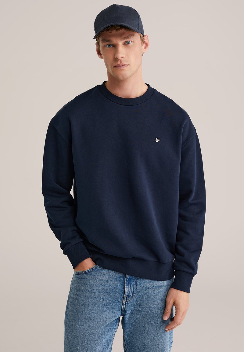 Temno modra jopa z okroglim izrezom in rebrastimi manšetami, z majhnim logotipom na prsih. Kombinirana s svetlo modrimi kavbojkami iz jeansa.