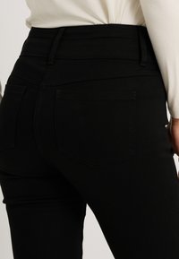 Jean noir taille haute avec une texture lisse, doté de deux poches arrière et de coutures minimales le long de la ceinture et des poches.