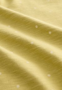 Tessuto giallo chiaro con una texture morbida, caratterizzato da piccoli puntini bianchi sparsi in un motivo regolare, drappeggiato per mostrarne il flusso.