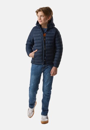 Junge trägt eine marineblaue Steppjacke, blaue Jeans und weiße Turnschuhe, steht mit einer Hand in der Tasche und schaut zur Seite.