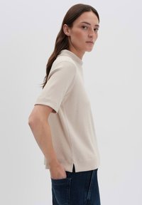 Donna in cima a manica corta a coste beige e jeans blu scuro con una mano in tasca, di profilo contro uno sfondo chiaro e uniforme.