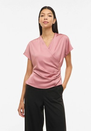 Kobieta o długich ciemnych włosach, ubrana w satynową, mauve bluzkę zapinaną na kopertę oraz czarne spodnie, stojąca z jedną ręką w kieszeni na białym tle.