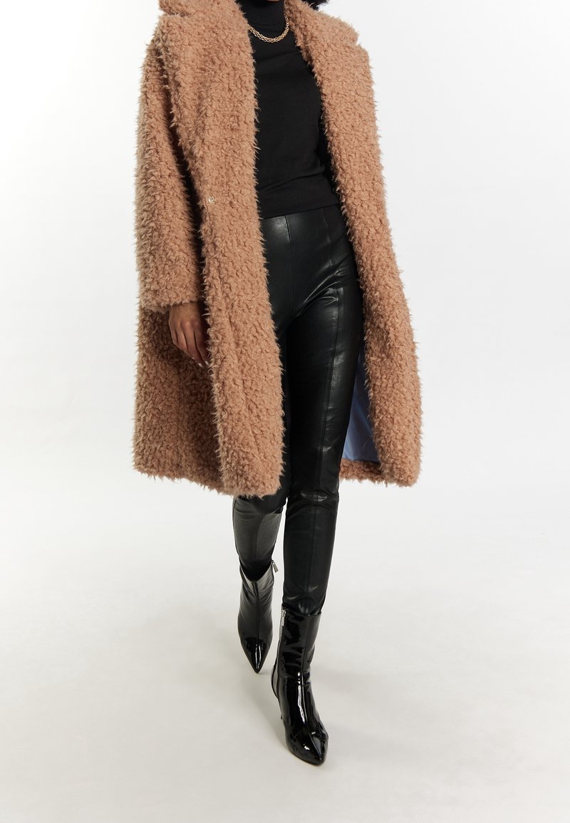 Femme portant un manteau en fausse fourrure beige, un col roulé noir, un pantalon en cuir noir et des bottines noires vernies à bout pointu, marchant vers l'avant.