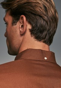 Camisa marrón con un cuello estructurado y un detalle de botón decorativo en la parte posterior. Textura suave con un sutil brillo, diseñada para un ajuste entallado.