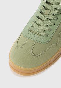 Világoszöld sneaker texturált szövet felsőrésszel, barna gumitalppal és azonos színű fűzőkkel. Díszített varrásokkal és sima orral rendelkezik.