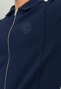 Veste zippée bleu marine en tissu texturé, avec un petit logo en relief sur la poitrine et un design de col classique.