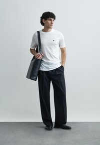 Bílé bavlněné tričko s malým černým logem, doplněné černými plisovanými kalhotami a velkou černou koženou kabelkou přes rameno. Celý vzhled zdobí hladké textury.